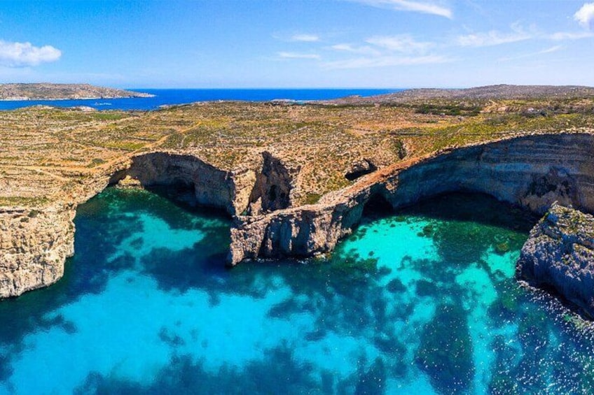 A coastal landscape with a prominent rock formation arching over a turquoise sea, surrounded by a hilly terrain. 2