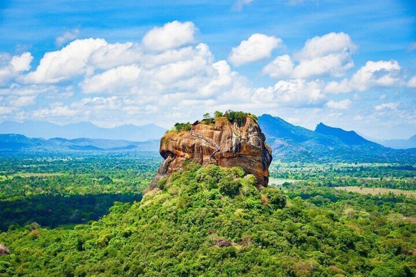 A large rock formation with a small peak, surrounded by dense green forest and mountains in the background. 1