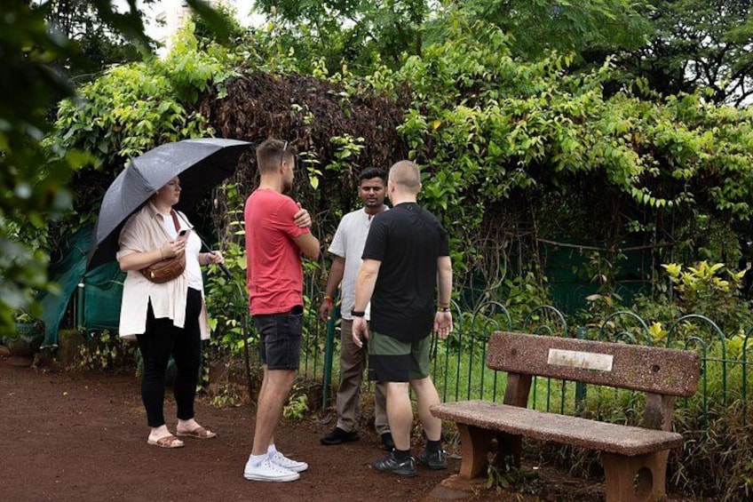Een groep mensen staat op een onverhard pad in een park met groen en een stenen bank. 2