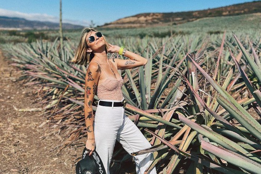 A person in a field with agave plants, wearing a pink top and white pants, with tattoos on their arms. 3