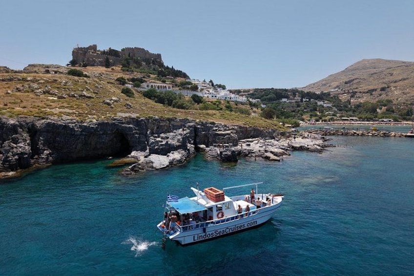 A boat named 'LindosSeaCruises.com' on a clear blue sea with rocky coastlines and a castle in the background. 2