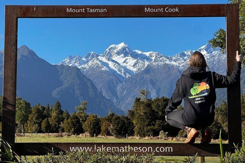 En person med en svart huvtröja med ett färgstarkt motiv, som står framför en inramad bild av bergen Mount Tasman och Mount Cook. 2