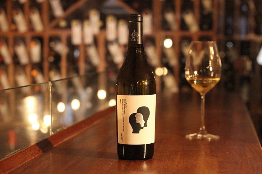 A wine bottle with a label featuring a silhouette of a person, placed on a wooden bar counter with a glass of white wine beside it. 2