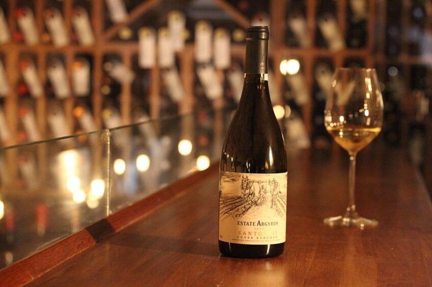 A bottle of ESTATE ARGYROS SANTO VINO 2011 wine on a wooden bar counter with a glass of white wine beside it. 3