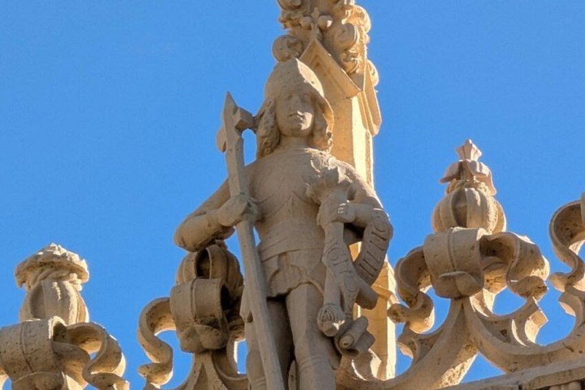 A detailed stone sculpture with intricate carvings, including a figure holding a staff and a shield, set against a clear blue sky. 3