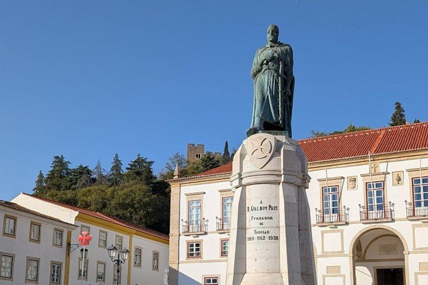 A statue on a pedestal with inscriptions, a building with a red roof, and a clear blue sky. 4