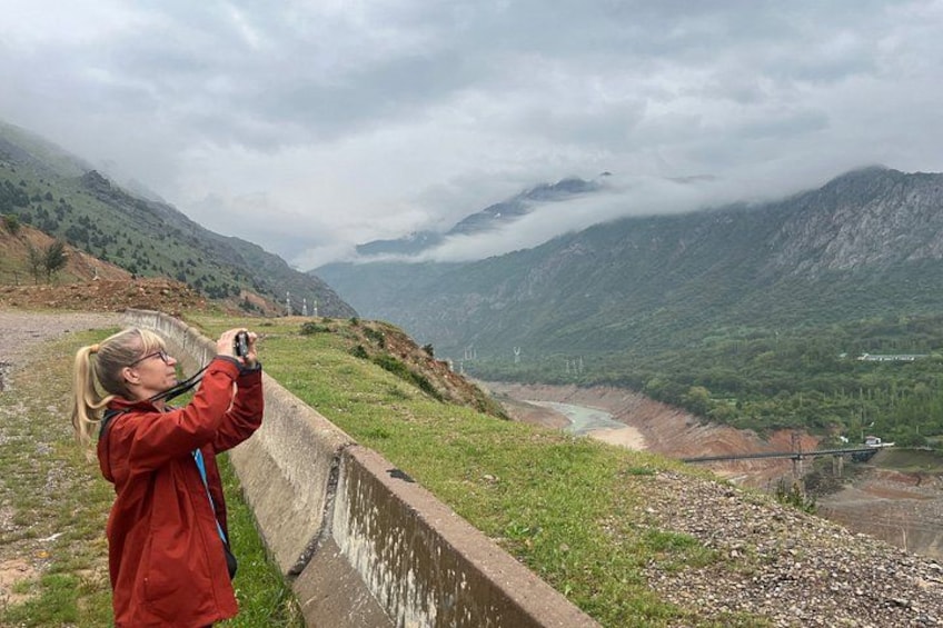 A person taking a photo of a mountainous landscape with a river and a bridge. 3