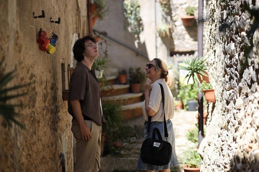 Two people standing in a narrow alleyway with potted plants and a textured wall. 2