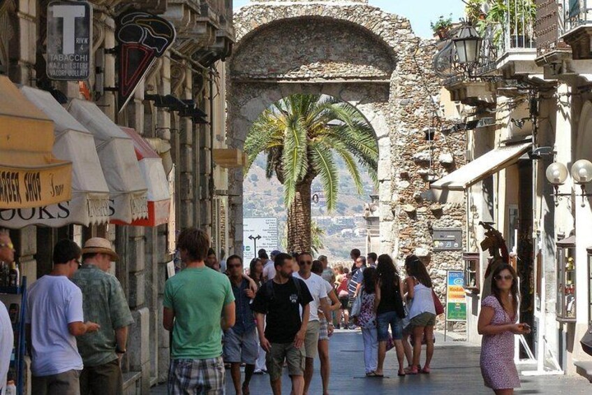 Une rue animée, où l’on voit des passants, un palmier et des bâtiments en pierre. 2