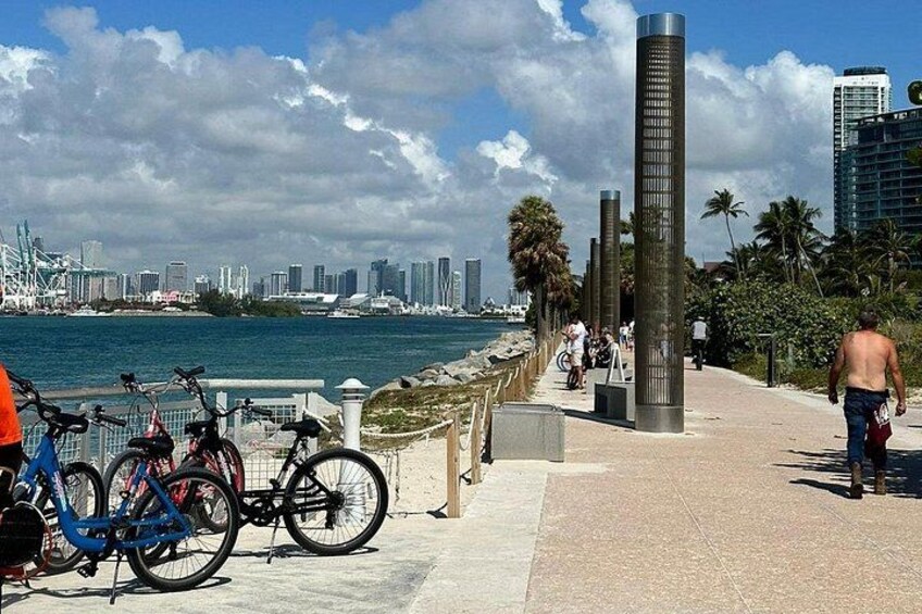 A waterfront promenade with bicycles, a tall sculpture, and a city skyline in the background. 2