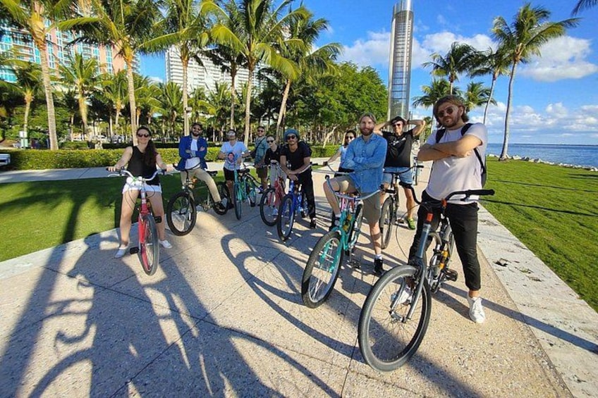 A group of people on bicycles along a paved path with palm trees and a tall building in the background. 1