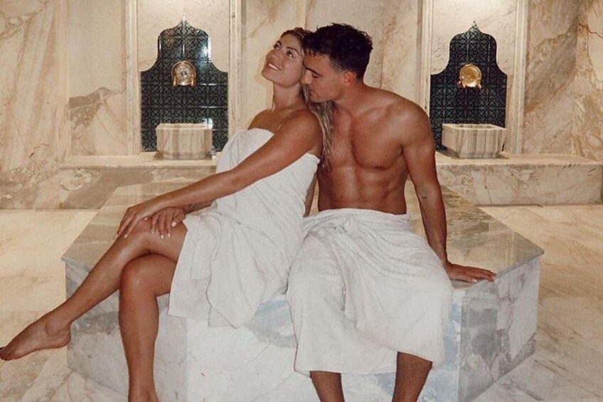 A marble bathroom with two people sitting on a bench, one with a towel wrapped around their body. 4