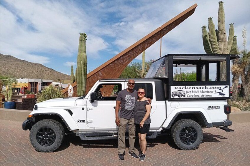 Un Jeep blanc, doté d’un porte-bagages noir, est garé dans une zone désertique, avec des cactus et une structure en bois en arrière-plan. 2