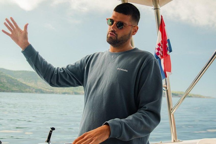 A person standing on a boat with a flag, water, and hills in the background. 2