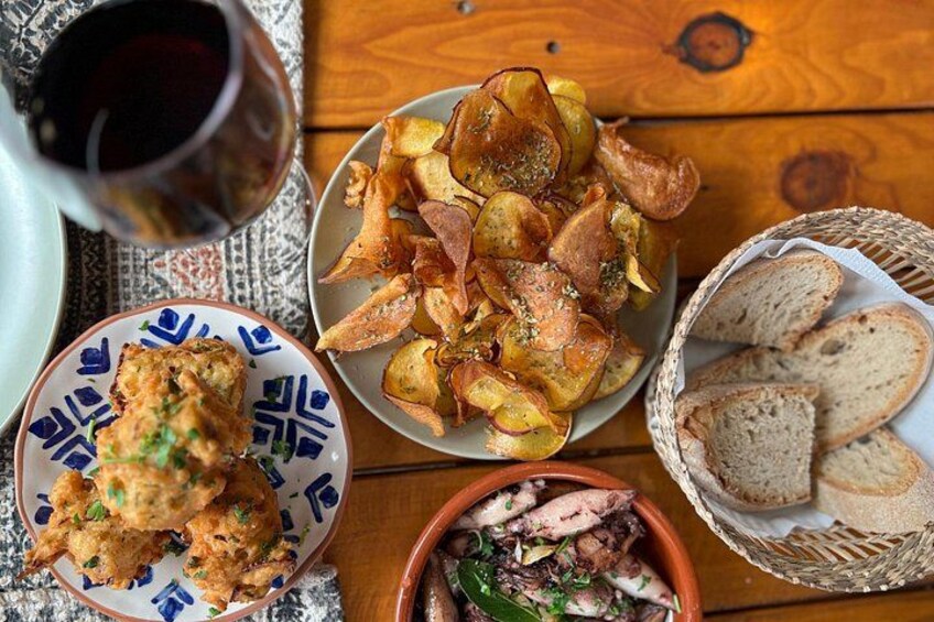 A wooden table with a plate of fried fish, a bowl of roasted potatoes, a basket of bread, and a glass of red wine. 1
