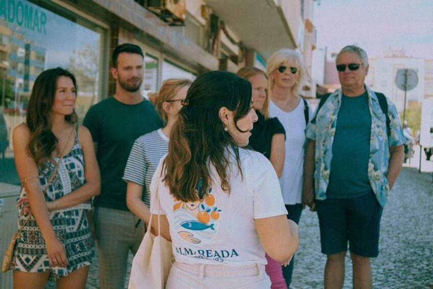 A group of people standing on a cobblestone street, with a building labeled 'ADOMAR' in the background. 3