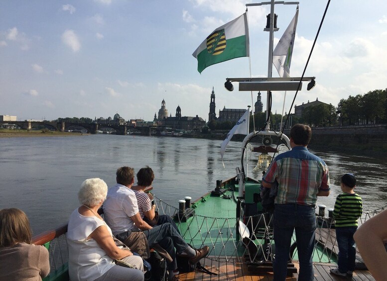A group of people on a boat with a flag, a river, and a cityscape with buildings and a bridge in the background. 4