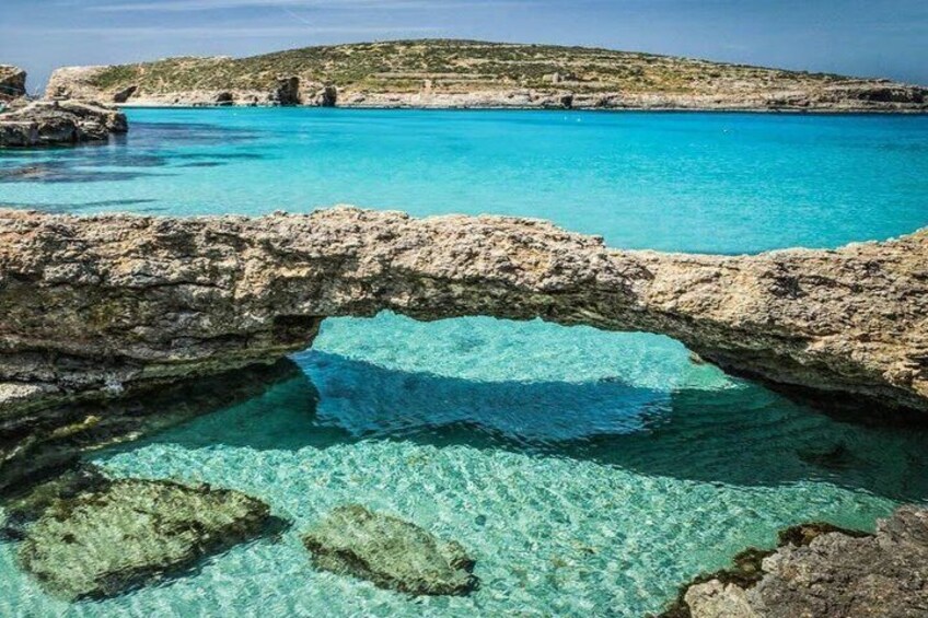 A coastal landscape with clear turquoise waters, a rocky arch formation, and distant hills. 1