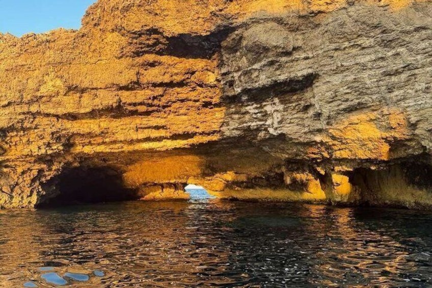 Un acantilado rocoso con la entrada de una cueva y agua tranquila que refleja el acantilado. 2