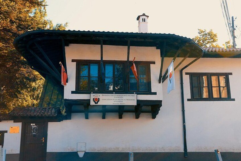 Ein Gebäude mit weißer Fassade, schwarzer Verzierung und einem Ziegeldach. An der Fassade hängen Flaggen, und ein Schild mit Text ist sichtbar. 4