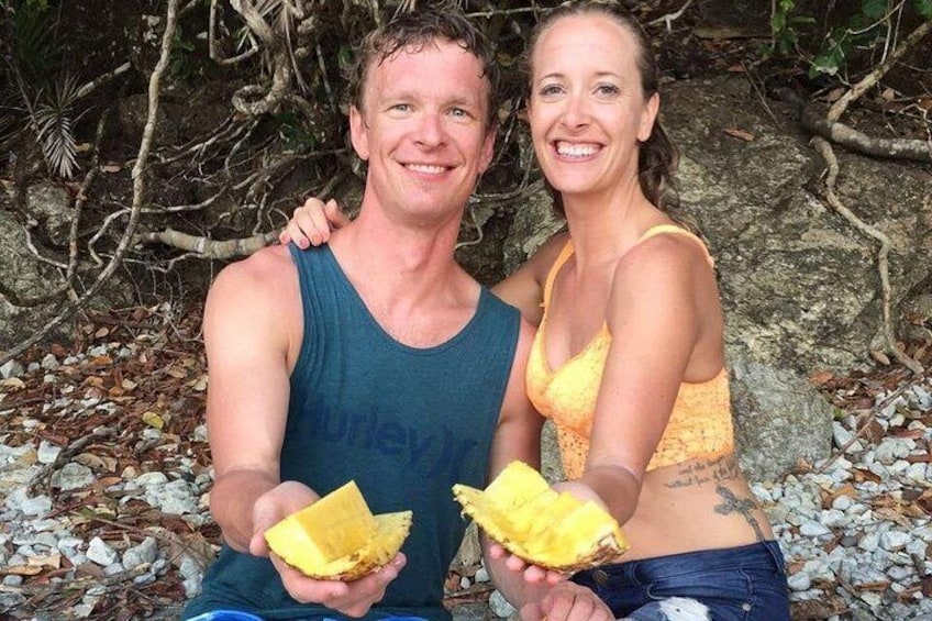 Two people holding pieces of fruit in a natural setting with rocks and foliage. 2