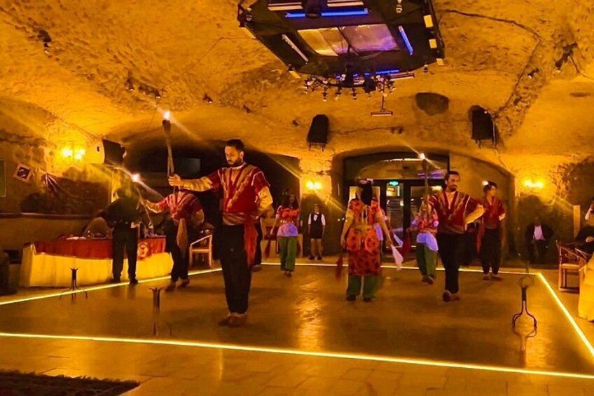 A group of people performing a traditional dance in a dimly lit, cave-like venue with a wooden floor and stone walls. 4