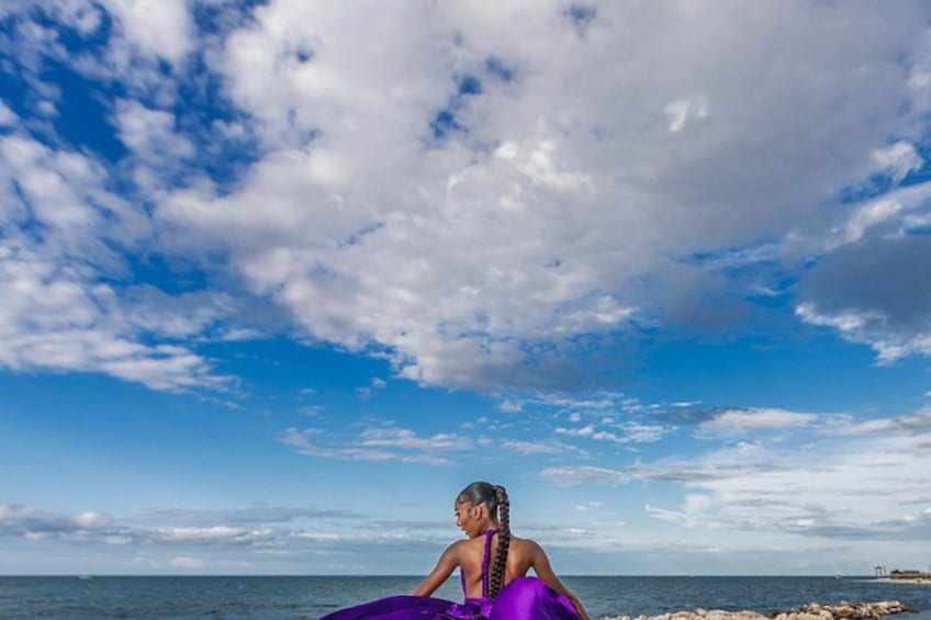 Una persona sentada en la playa, vestida con un vestido morado, mirando hacia el océano bajo un cielo parcialmente nublado. 2