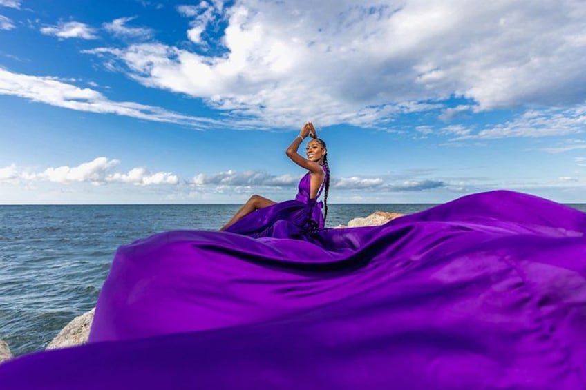 Una persona con un vestido morado sentada sobre una roca junto al mar, bajo un cielo despejado. 1
