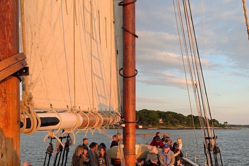 A sailboat with a group of people on deck, a large wooden mast, and a calm body of water with a distant shoreline. 5