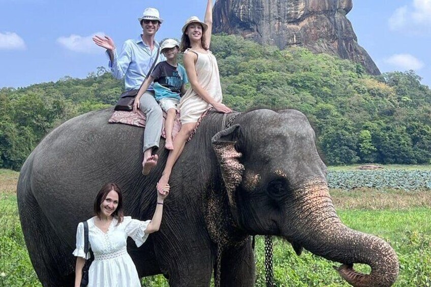 Une famille chevauche un éléphant dans un champ herbeux, avec une grande formation rocheuse en arrière-plan. 2