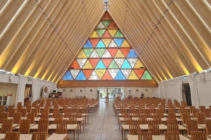 A large hall with rows of wooden chairs and tables, a triangular stained glass window, and a high, curved ceiling. 3