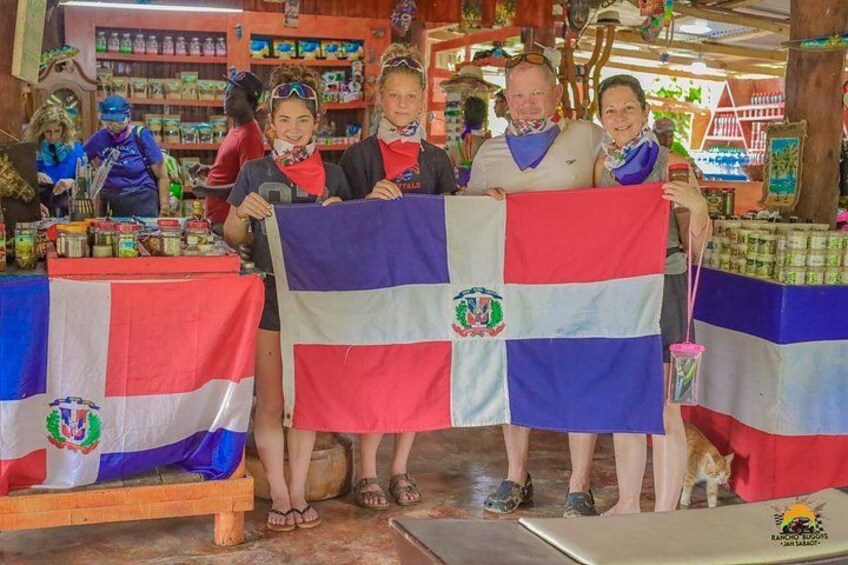 Un grupo de personas sostiene una bandera con un escudo, situadas en un mercado con estantes llenos de productos y una mesa de madera. 5