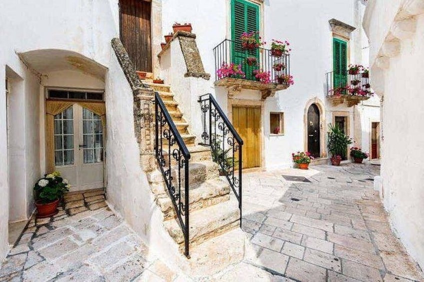 A narrow stone-paved alley with a staircase leading to a building with green shutters and a balcony adorned with flowers. 2