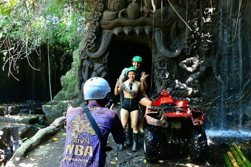 A person in a purple jacket with 'NBA' and 'Adventure' on it, a red ATV, and a cave entrance with intricate carvings. 1