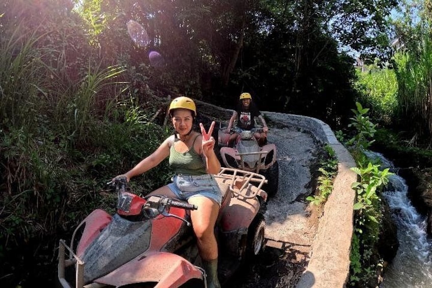 Two people on quad bikes in a narrow water channel surrounded by greenery. 3