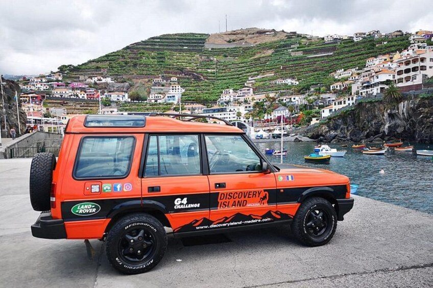 An orange Land Rover G4 Discovery Islander parked by a waterfront with a coastal village in the background. 2