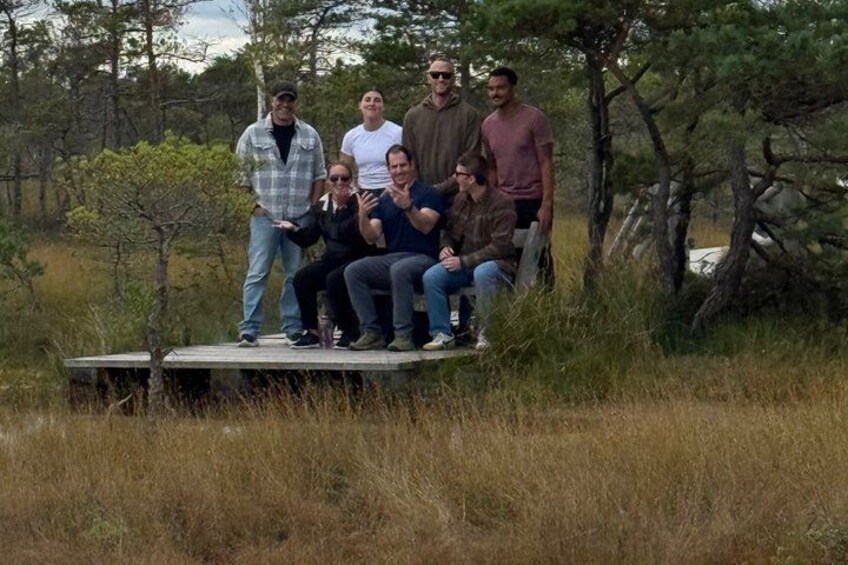 Eine Gruppe Menschen auf einer Holzplattform in einem grünen Feld, im Hintergrund Bäume. 2