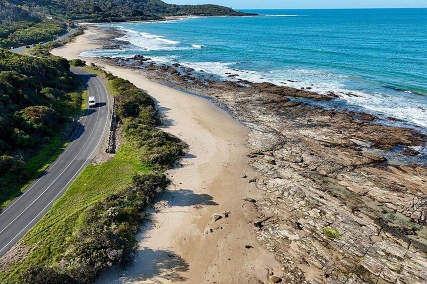 A coastal road with a vehicle, beach, and rocky shoreline. 1