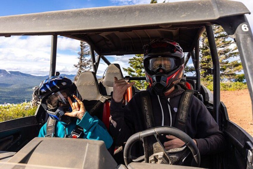 Two people in a golf cart, one driving and one passenger, with a mountainous landscape in the background. 3