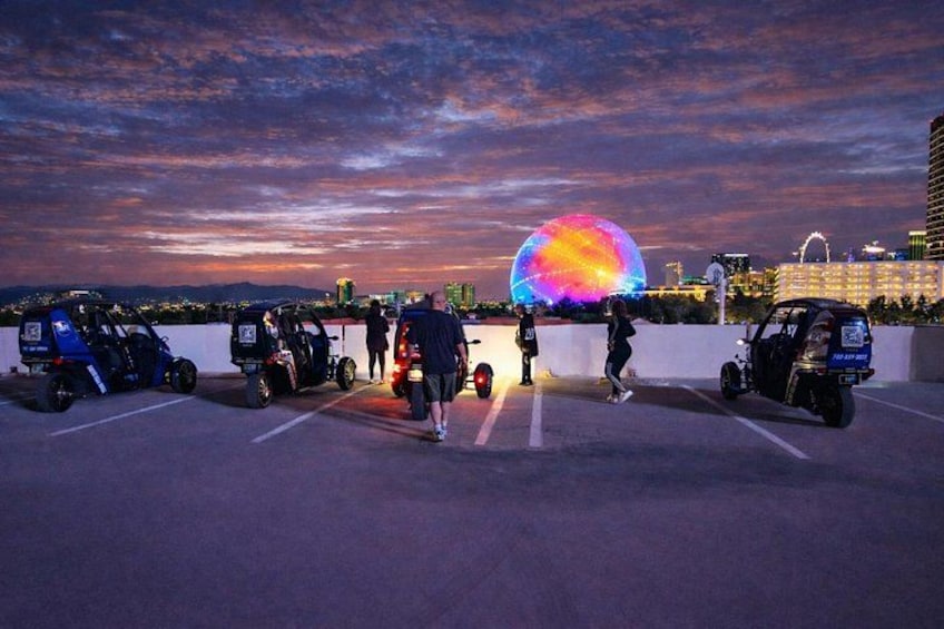 Un estacionamiento con patinetas eléctricas, un gran globo iluminado y el horizonte de una ciudad al atardecer. 3