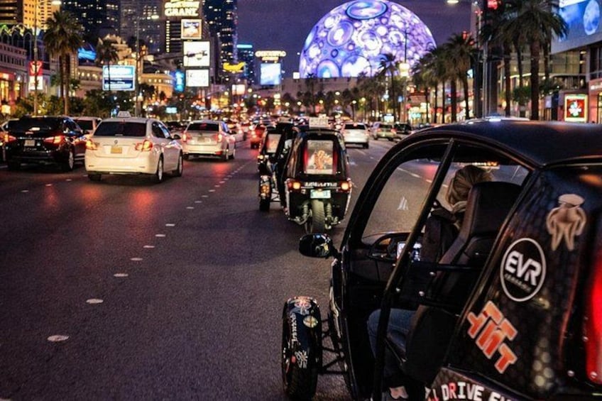 Una concurrida calle urbana de noche con varios vehículos, incluyendo automóviles y un tuk-tuk, y vallas publicitarias iluminadas. 4