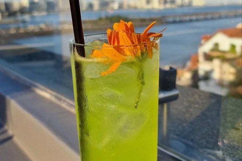 A tall glass with a green drink, ice cubes, and orange flower garnish, viewed from a high vantage point overlooking a body of water and buildings. 4