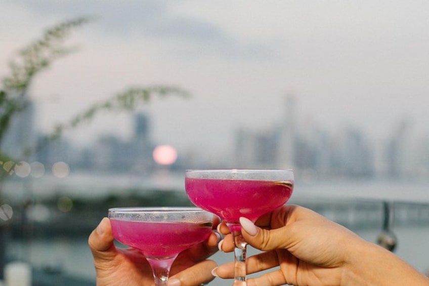Two hands holding martini glasses with pink drinks, a blurred city skyline in the background, and a body of water. 3