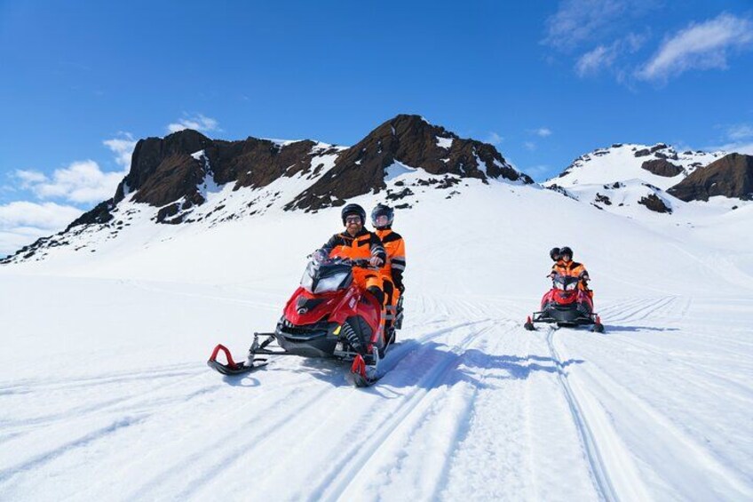 Zwei Personen fahren mit Schneemobil auf einem verschneiten Berg bei strahlendem Himmel. 2