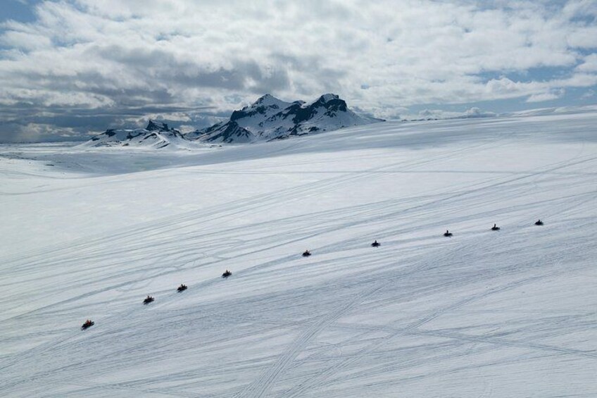 Schneebedeckte Landschaft, in der eine Fahrzeuggruppe einen Hang hinabfährt, dahinter ein Gebirge und ein teilweise bewölkter Himmel. 5