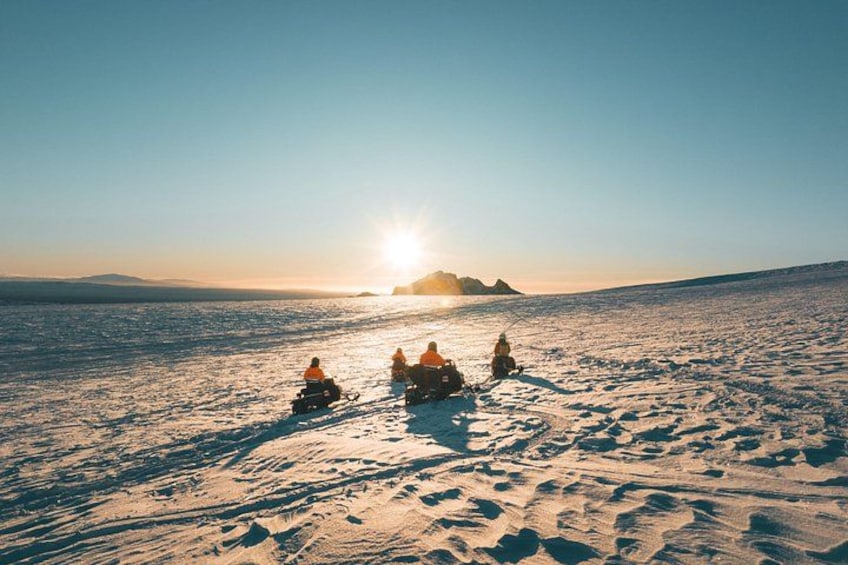 Eine Gruppe von Personen fährt mit Schneemotoren durch eine verschneite Landschaft, im Hintergrund versinkt die Sonne. 4