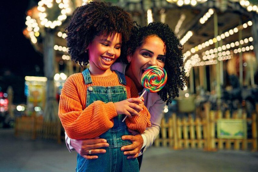 Two people embracing, one holding a lollipop, in a brightly lit outdoor setting with decorative lights. 3