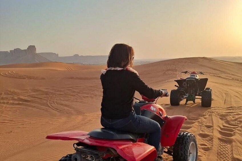 A person riding a red ATV on a desert trail with sand dunes and a distant rocky formation. 3
