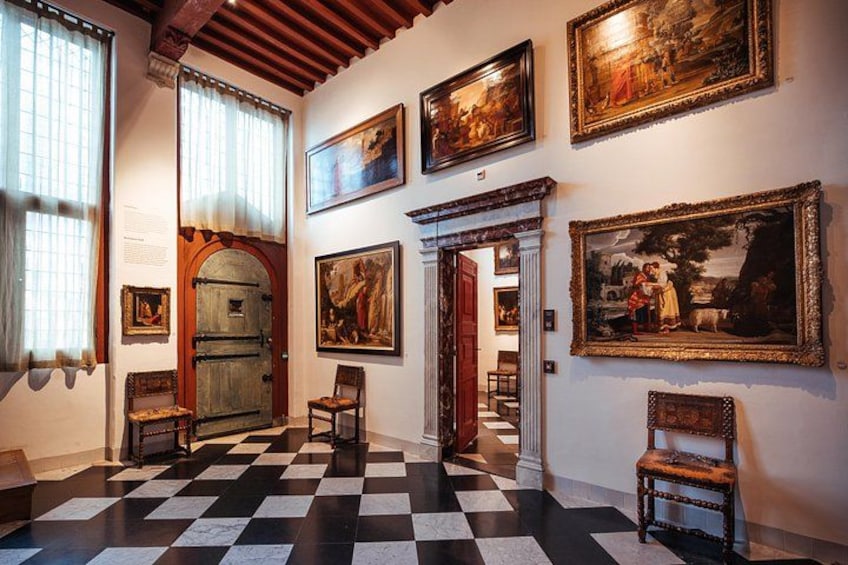 A hallway with a checkered floor, wooden doors, and framed paintings on the walls. 4