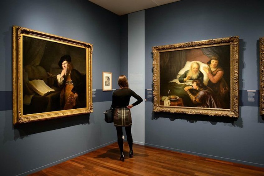 Interno di un museo con due grandi dipinti appesi alle pareti e una persona che li osserva. 1